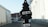 A black, tree-shaped car air freshener labeled 'Little Trees' and 'Black Ice' is hanging in front of a blurred outdoor background. The setting appears to be an alley or street with buildings on either side.
