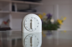 A white kitchen timer is placed on a reflective surface, showing a clear reflection below it. The timer is circular with numbers around the edge and a knob in the center. In the blurred background, colorful flowers add a touch of vibrancy.