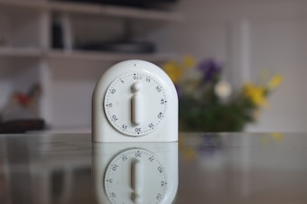 A white kitchen timer is placed on a reflective surface, showing a clear reflection below it. The timer is circular with numbers around the edge and a knob in the center. In the blurred background, colorful flowers add a touch of vibrancy.