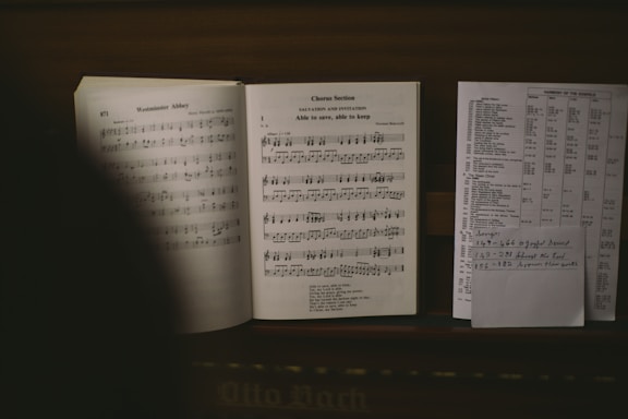 An open music book with visible musical notes and lyrics lies on top of a wooden surface. Next to it, there is a sheet of paper with text and a handwritten note listing song titles and numbers.
