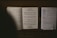 An open music book with visible musical notes and lyrics lies on top of a wooden surface. Next to it, there is a sheet of paper with text and a handwritten note listing song titles and numbers.