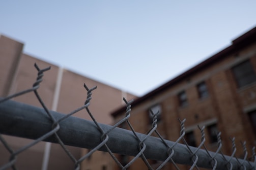 A close-up view of a chain-link fence with twisted barbed wire on top, set against the backdrop of a building with brick walls and windows. The image has a clear sky in the background.
