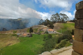 A lush, hilly landscape features several rustic buildings with red tile roofs nestled among vibrant green fields. Trees rise on the hillsides, with some areas appearing slightly smoky, possibly from burning vegetation or mist. In the foreground, part of an ancient stone structure is visible.