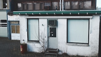 An old, weathered building facade with a white exterior and green roof trim. Two large windows with vertical blinds flank a central door marked with a sign. A bare tree stands in front of the building, and a rusted barrel sits nearby.