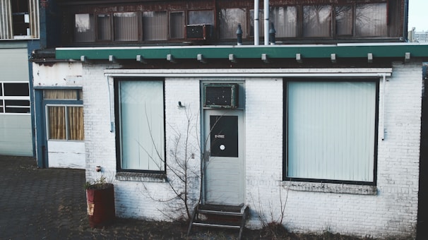 An old, weathered building facade with a white exterior and green roof trim. Two large windows with vertical blinds flank a central door marked with a sign. A bare tree stands in front of the building, and a rusted barrel sits nearby.