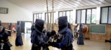 A group of people engaged in kendo practice inside a spacious gymnasium. Two individuals are in the foreground wearing traditional kendo uniforms and helmets, holding bamboo swords. The room has wooden walls, a chalkboard, and large windows letting in natural light.