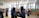 A group of people engaged in kendo practice inside a spacious gymnasium. Two individuals are in the foreground wearing traditional kendo uniforms and helmets, holding bamboo swords. The room has wooden walls, a chalkboard, and large windows letting in natural light.