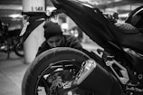 A black and white image of a person kneeling beside a motorcycle, closely inspecting the rear wheel and exhaust. The setting appears to be an indoor garage with other vehicles in the background, indicated by the concrete pillars and dim lighting.