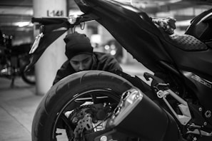 A black and white image of a person kneeling beside a motorcycle, closely inspecting the rear wheel and exhaust. The setting appears to be an indoor garage with other vehicles in the background, indicated by the concrete pillars and dim lighting.
