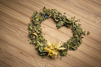 A heart-shaped arrangement of dried leaves and flowers lies on a wooden surface. The dried plants include green leaves and petals of various colors, such as yellow and blue, with a prominent yellow flower at the bottom of the heart shape.