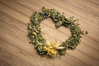 A heart-shaped arrangement of dried leaves and flowers lies on a wooden surface. The dried plants include green leaves and petals of various colors, such as yellow and blue, with a prominent yellow flower at the bottom of the heart shape.