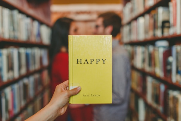A yellow book with the title 'HAPPY' by Alex Lemon is held in the foreground with a library aisle in the background. A blurred view of two people, one wearing a red top, standing and facing each other amidst the bookshelves.