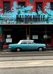 A vintage turquoise car is parked in front of a building featuring vibrant graffiti art and the word 'PALOOKAVILLE'. The building has a red and turquoise color scheme with murals depicting abstract figures. Signs indicating 'For Lease' are visible in the windows.
