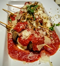 A dish featuring skewered meatballs covered in tomato sauce and garnished with grated cheese and fresh basil. The presentation is on a rectangular white plate, with a sprinkle of herbs and seasoning on top.