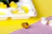 Small rubber ducks in yellow and white are arranged on a striped purple and white surface. Some ducks are placed on a white tray with compartments filled with yellow liquid. The background consists of vibrant yellow and purple colors.