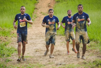 Four men covered in mud are running on a dirt path surrounded by lush green grass. They are wearing blue shirts and athletic shoes, suggesting participation in an outdoor obstacle or mud race.