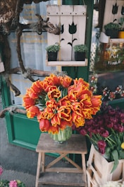 A vibrant floral arrangement of tulips with red petals and yellow fringes sits in a glass vase on a wooden stool. The background includes a green painted door adorned with plant holders and decorative wood pieces. More flowers and small potted plants surround the area, contributing to a rustic, garden shop ambiance.