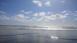 A serene ocean view with gentle waves rolling towards the shore. The sky is bright and filled with scattered fluffy clouds, reflecting sunlight that shimmers off the water's surface.