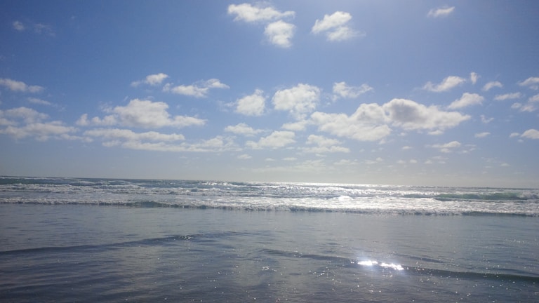 A serene ocean view with gentle waves rolling towards the shore. The sky is bright and filled with scattered fluffy clouds, reflecting sunlight that shimmers off the water's surface.
