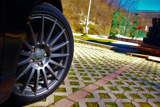 A close-up of a car wheel with a visible logo, positioned over a patterned pavement with grass between the tiles. In the background, there are stacks of materials and a green metal structure, set against a backdrop of trees and a bright blue sky.