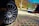 A close-up of a car wheel with a visible logo, positioned over a patterned pavement with grass between the tiles. In the background, there are stacks of materials and a green metal structure, set against a backdrop of trees and a bright blue sky.