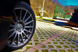 A close-up of a car wheel with a visible logo, positioned over a patterned pavement with grass between the tiles. In the background, there are stacks of materials and a green metal structure, set against a backdrop of trees and a bright blue sky.
