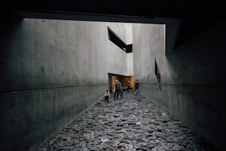 A long, narrow corridor with gray, concrete walls creates a stark and austere atmosphere. The ground is covered with metal disc-shaped objects, each with cut-out facial expressions. People, including a child, walk through the space, adding a sense of scale and interaction within the somber setting.