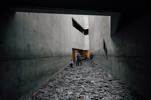 A long, narrow corridor with gray, concrete walls creates a stark and austere atmosphere. The ground is covered with metal disc-shaped objects, each with cut-out facial expressions. People, including a child, walk through the space, adding a sense of scale and interaction within the somber setting.