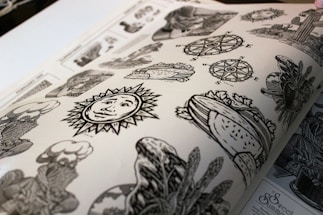 A page from a book featuring various black and white illustration designs, including a sun with a face, compass roses, a heart with foliage, and assorted decorative motifs. The artwork is detailed and appears to be printed on matte paper.