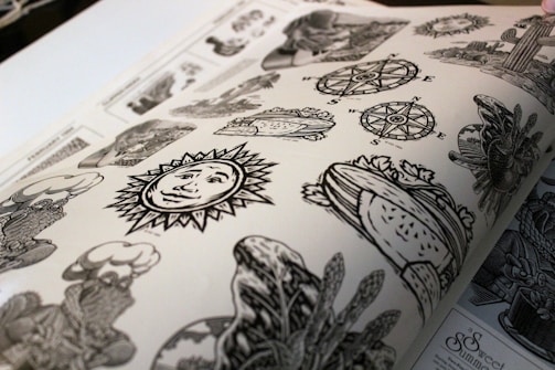 A page from a book featuring various black and white illustration designs, including a sun with a face, compass roses, a heart with foliage, and assorted decorative motifs. The artwork is detailed and appears to be printed on matte paper.