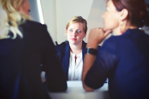 Three people are sitting at a table, engaged in a discussion. Two individuals are facing away from the camera, while one person is facing the camera attentively, appearing to listen intently. The setting appears professional, likely a meeting or interview.