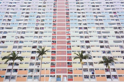 A large residential apartment building has a pastel-colored facade with a variety of windows and balconies. The structure features vertical rows of different colored panels, including shades of blue, yellow, pink, and red. In front of the building are several palm trees that add a touch of greenery to the urban setting.