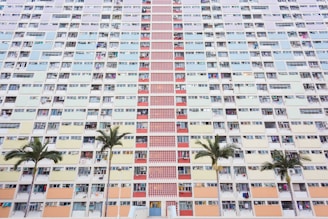 A large residential apartment building has a pastel-colored facade with a variety of windows and balconies. The structure features vertical rows of different colored panels, including shades of blue, yellow, pink, and red. In front of the building are several palm trees that add a touch of greenery to the urban setting.