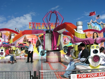 Amusement park ride with vibrant colors and several people sitting in seats attached to mechanical arms. A person wearing a white shirt is walking on the platform near the ride. The letters spelling 'Experience' are visible in the background above the ride, with colorful bulb lights decorating the structure. The blue sky and flags fluttering in the background add to the lively ambiance.