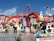 Amusement park ride with vibrant colors and several people sitting in seats attached to mechanical arms. A person wearing a white shirt is walking on the platform near the ride. The letters spelling 'Experience' are visible in the background above the ride, with colorful bulb lights decorating the structure. The blue sky and flags fluttering in the background add to the lively ambiance.