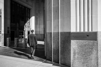 A monochrome image of a man in a suit walking towards the entrance of a building. The architecture features large stone pillars and rectangular windows, and a sign on the right reads 'Credit Suisse'. The lighting creates strong shadows on the ground.