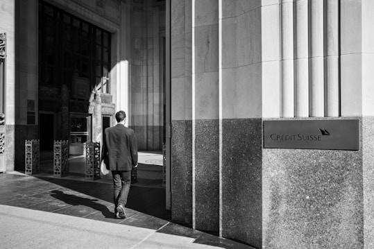 A monochrome image of a man in a suit walking towards the entrance of a building. The architecture features large stone pillars and rectangular windows, and a sign on the right reads 'Credit Suisse'. The lighting creates strong shadows on the ground.