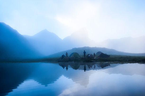 A tranquil landscape featuring a serene lake reflecting the silhouettes of distant mist-shrouded mountains. Several tents are pitched on the grassy shore, with a group of people gathered nearby, enjoying the peaceful natural setting.