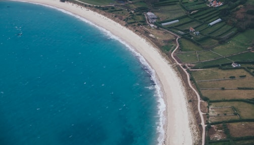 An aerial view of a coastline with clear turquoise water meeting a sandy beach. Adjacent to the beach is a patchwork of rectangular green fields, crisscrossed by pathways and bordered by hedges. Some small buildings and greenhouses are scattered across the landscape.