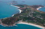 An aerial view of an island with curving white sandy beaches, surrounded by turquoise blue waters. The island features a patchwork of green agricultural fields and scattered buildings, with a more concentrated settlement towards the center. The coastline is indented with a mix of rocky and sandy shores.