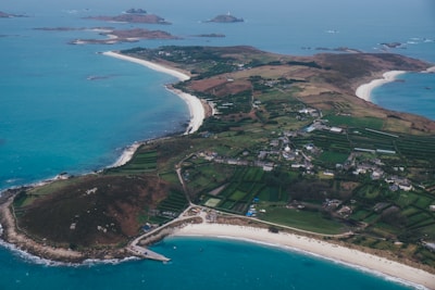 An aerial view of an island with curving white sandy beaches, surrounded by turquoise blue waters. The island features a patchwork of green agricultural fields and scattered buildings, with a more concentrated settlement towards the center. The coastline is indented with a mix of rocky and sandy shores.