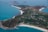 An aerial view of an island with curving white sandy beaches, surrounded by turquoise blue waters. The island features a patchwork of green agricultural fields and scattered buildings, with a more concentrated settlement towards the center. The coastline is indented with a mix of rocky and sandy shores.