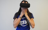 A person wearing a virtual reality headset covers their mouth with their hands, possibly expressing surprise or awe. They are dressed in a blue shirt with white lettering and have a tattoo on one arm, along with a colorful bracelet.