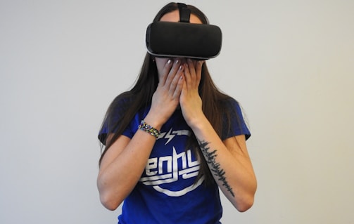 A person wearing a virtual reality headset covers their mouth with their hands, possibly expressing surprise or awe. They are dressed in a blue shirt with white lettering and have a tattoo on one arm, along with a colorful bracelet.