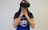 A person wearing a virtual reality headset covers their mouth with their hands, possibly expressing surprise or awe. They are dressed in a blue shirt with white lettering and have a tattoo on one arm, along with a colorful bracelet.