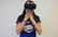 A person wearing a virtual reality headset covers their mouth with their hands, possibly expressing surprise or awe. They are dressed in a blue shirt with white lettering and have a tattoo on one arm, along with a colorful bracelet.