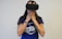A person wearing a virtual reality headset covers their mouth with their hands, possibly expressing surprise or awe. They are dressed in a blue shirt with white lettering and have a tattoo on one arm, along with a colorful bracelet.