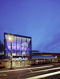A modern commercial building with large glass windows and illuminated signs at dusk. The signage reads 'Citi Plaza' with bright, white lights. The surrounding lighting creates a lively urban atmosphere with blurred light trails of passing vehicles on the street in front of the building.