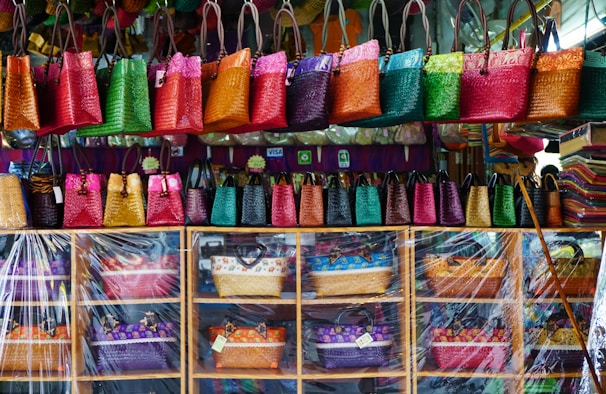 A colorful display of woven handbags arranged on shelves and hanging hooks. The bags come in vibrant shades of pink, green, orange, red, and yellow. There is a clear plastic sheet partially covering the lower shelves. The background includes a variety of additional textiles and a few payment method signs.