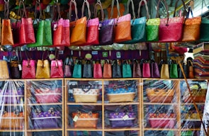 A colorful display of woven handbags arranged on shelves and hanging hooks. The bags come in vibrant shades of pink, green, orange, red, and yellow. There is a clear plastic sheet partially covering the lower shelves. The background includes a variety of additional textiles and a few payment method signs.